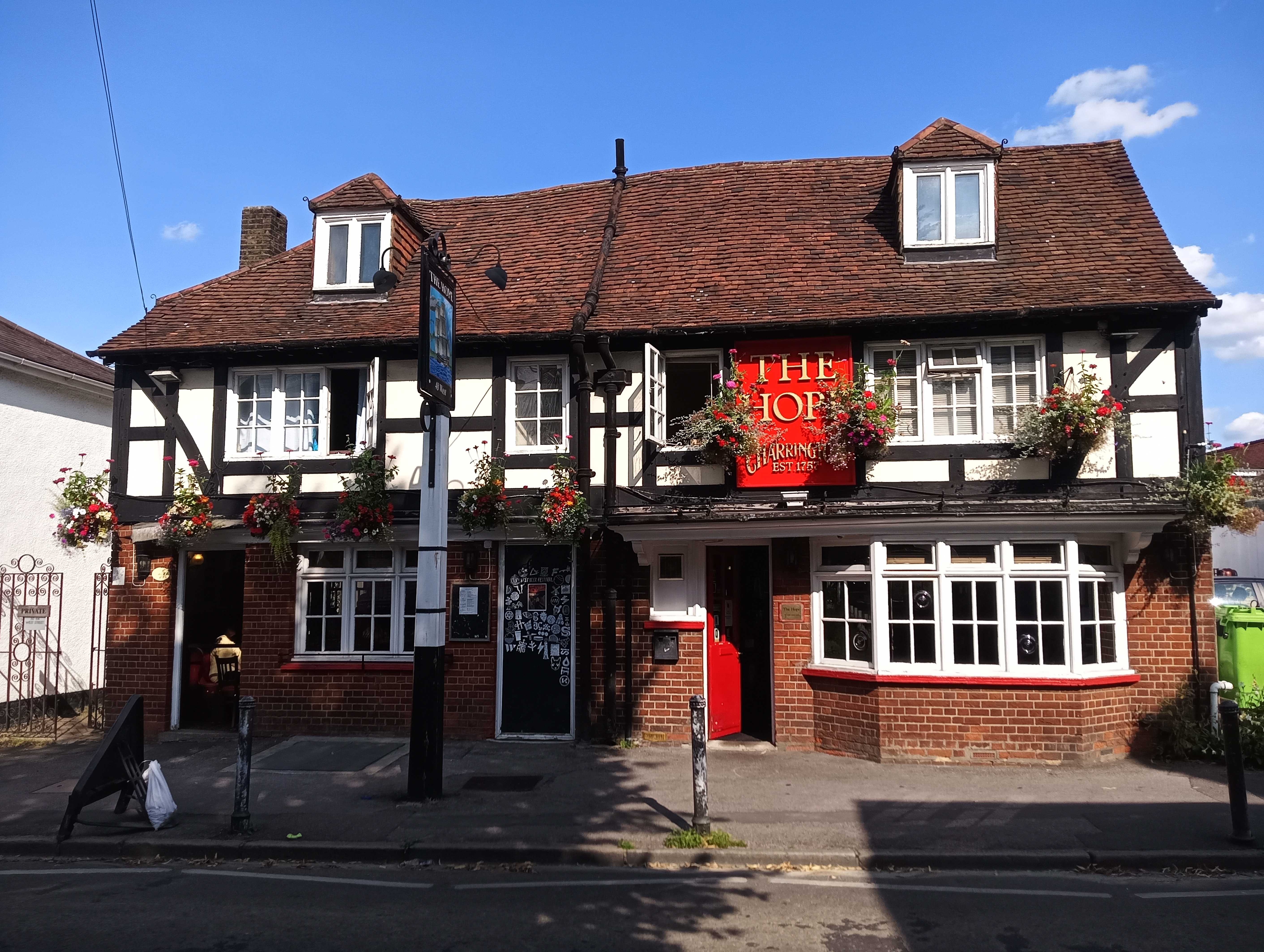 A photo of the Hope by Rodger Molyneux. A traditional looking pub building with redbrick and black timber framing with white paint. A red and gold sign is in the centre of the pub, above the red door. Hanging flower baskets are above the ground floor windows and a traditional pub sign is outside the pub, which features a painted image of a ship.