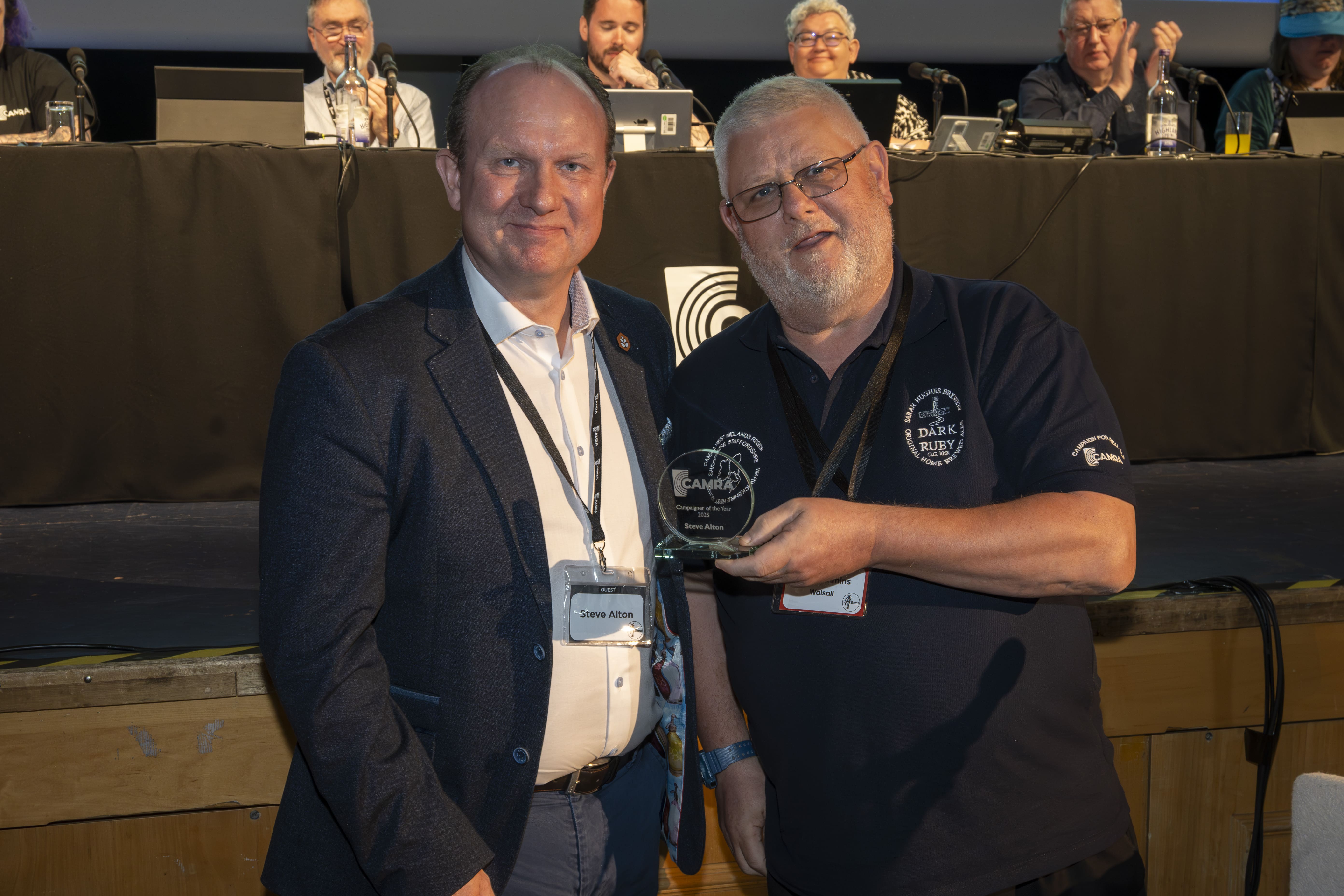Steve Alton (right) wearing a black suit and white shirt with a lanyard is present a small glass trophy by Gary Timmins, wearing a navy polo shirt. Behind them is a stage with a long table draped in black