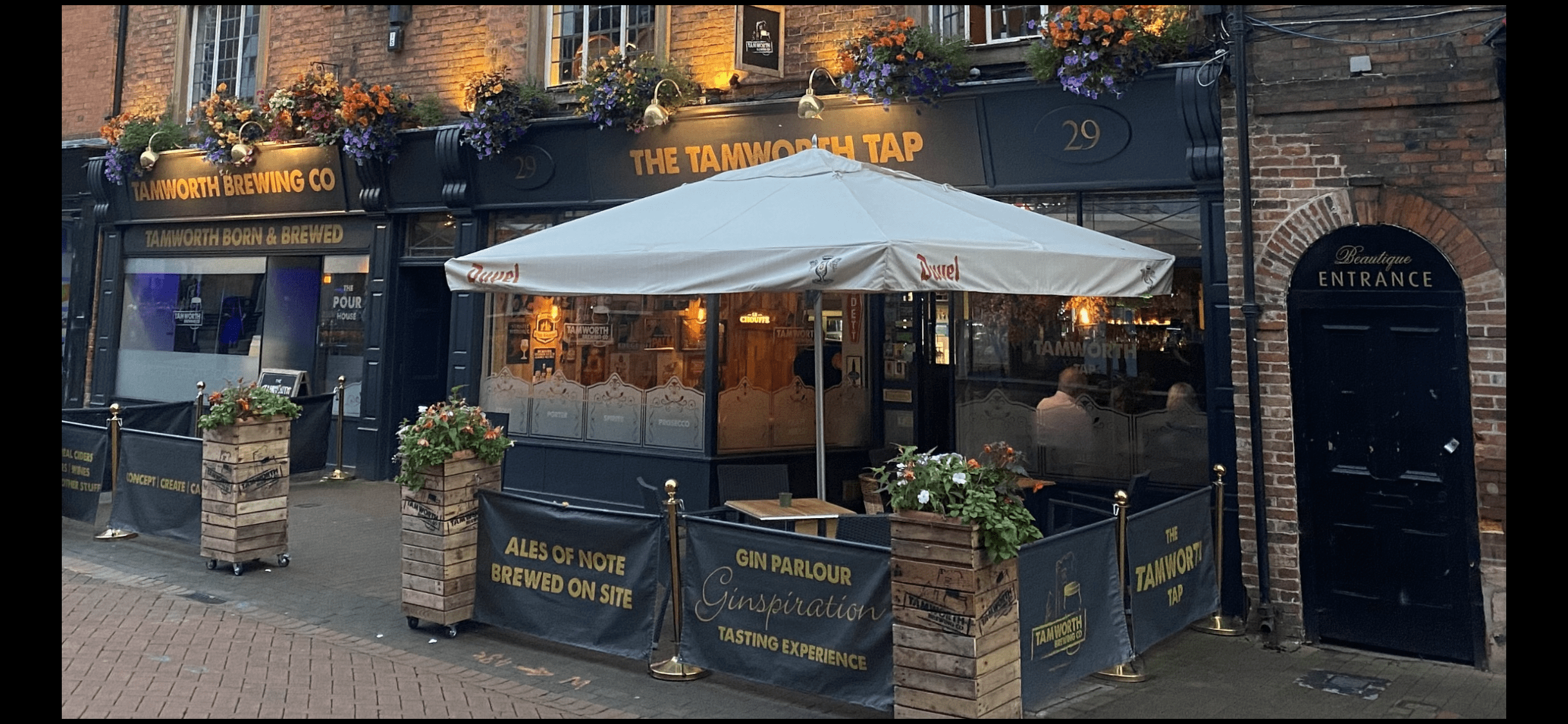 A large white parasol outside a black painted pub with gold lettering. Wooden flower cases are in front of the pub