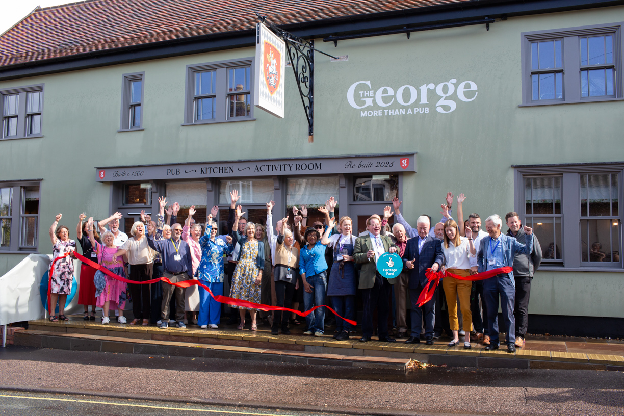 Photo of the exterior of the George, Wickham Market, with the committee members of the pub in front celebrating their official opening. The colour of the pub is mint green with a large hanging sign with the St George Coat of Arms on it.