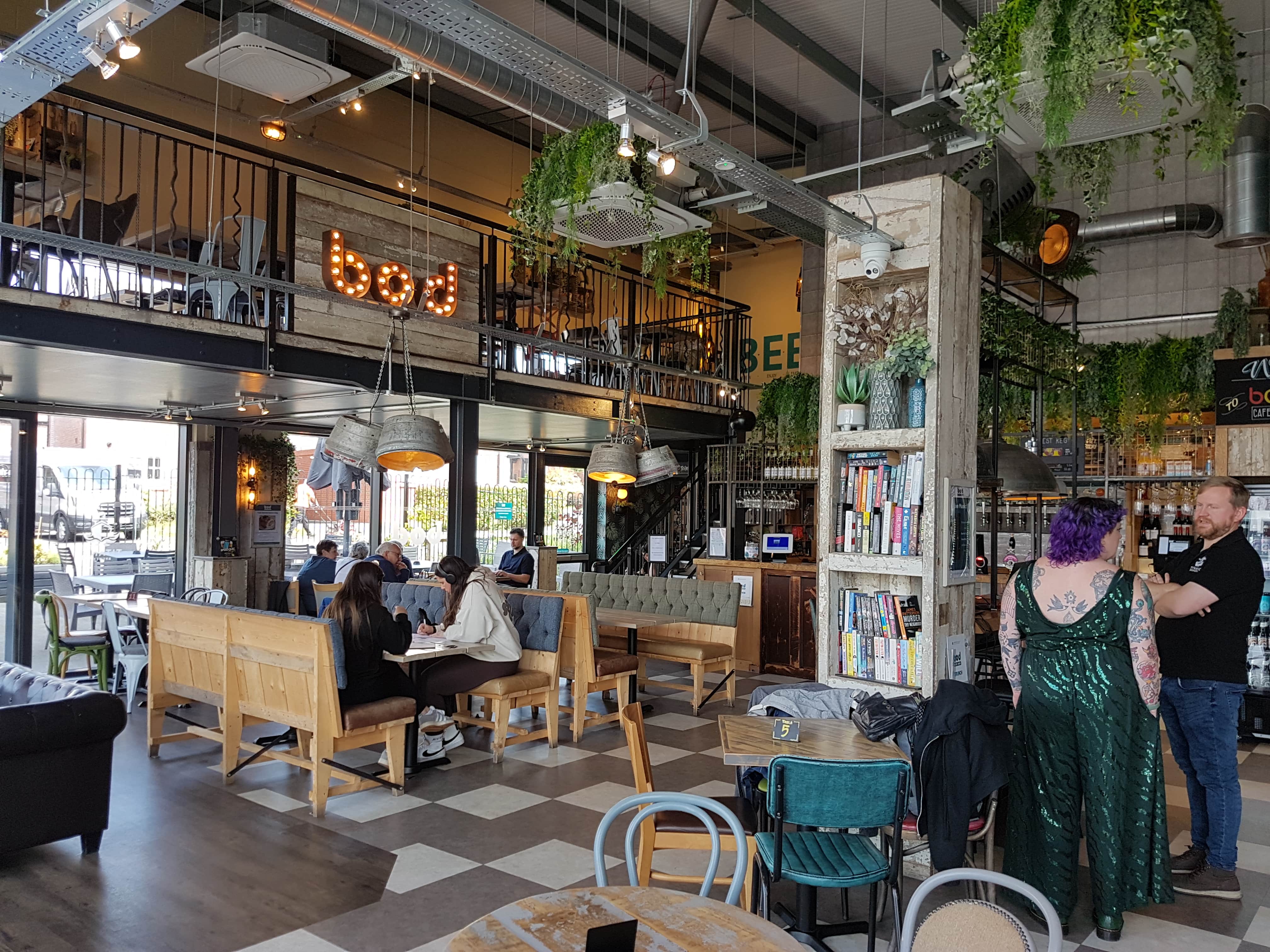 A large open space with checkered flooring, wooden panelling with distressed paintwork, hanging plants, light fittings made from steel barrels and exposed ceiling pipes. To the right there is a tall book case and in the middle is a group of people sitting down at a table with benches.