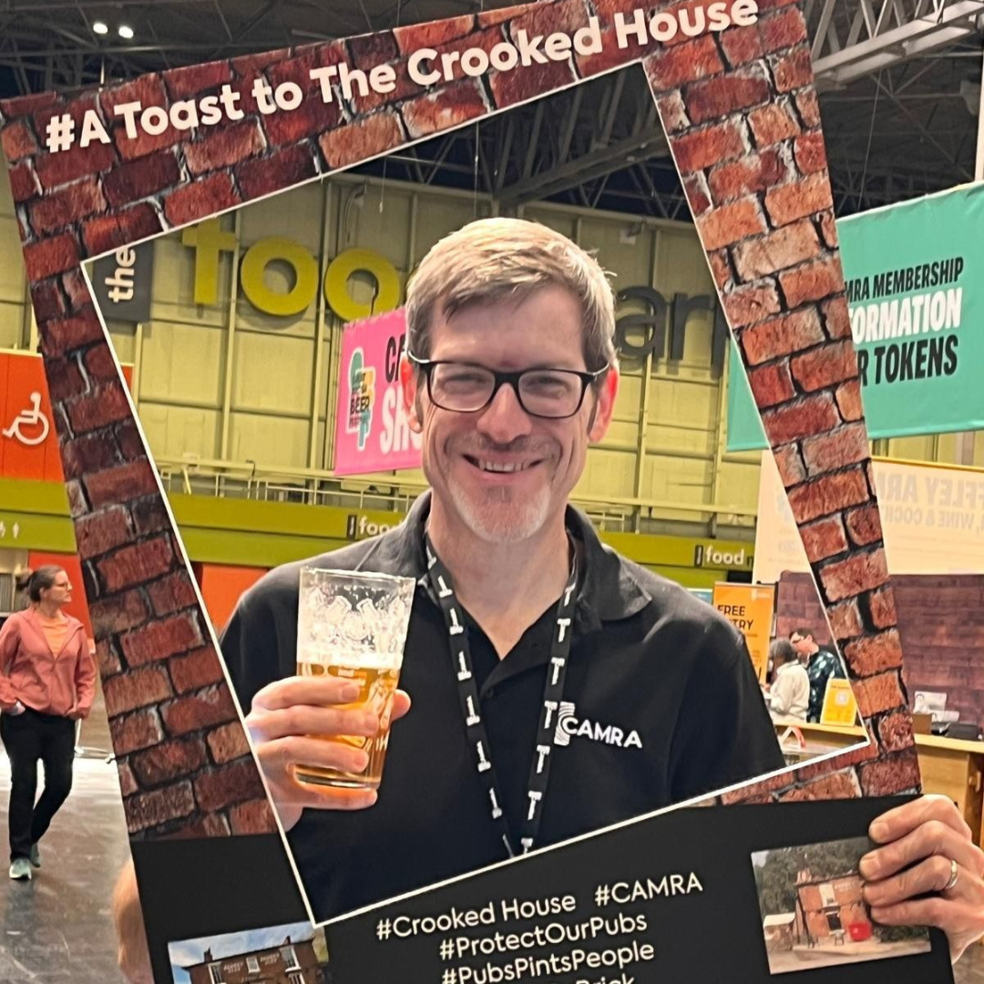 CAMRA Chief Executive Tom Stainer holding a selfie prop cardboard cut out for #ToastToTheCrookedHouse campaign. He is smiling and is holding a pint at Great British Beer Festival 2025.