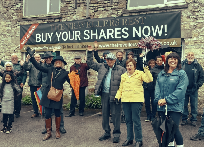 Photo of community members of Skeeby outside the closed Travellers Rest. They are over 20 adults and children posing for a photo, with a banner on the pub saying 'The Traveller's Rest, Buy Your Shares Now!'.