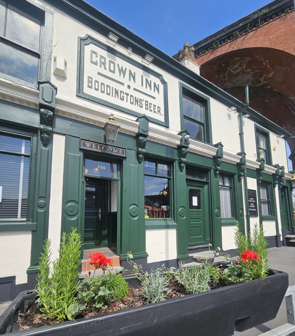 Photo of the exterior of the Crown Inn. In front of the pub is a long plant pot, with flowers in. The pub is cream and green colored, with its name and 'Boddingtons' Beer' on the front of the pub's wall.