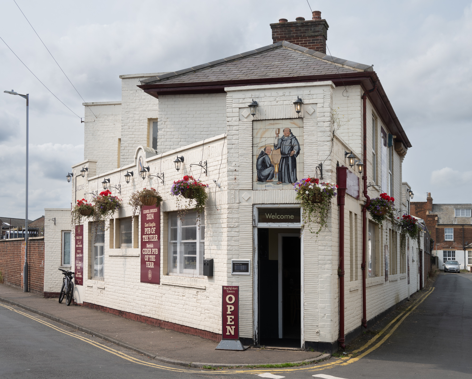 A white building with red signs between windows and hanging baskets. A decal of two monks is above the door