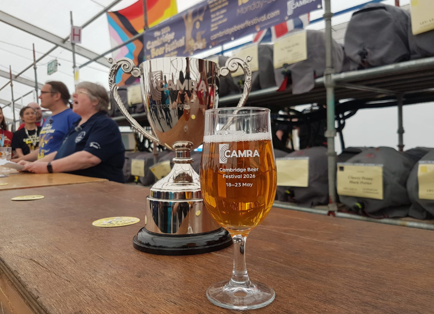 A shiny silver trophy and a clear glass of beer are placed on a wooden table at the Cambridge Beer Festival 2026. People and beer casks in the background.