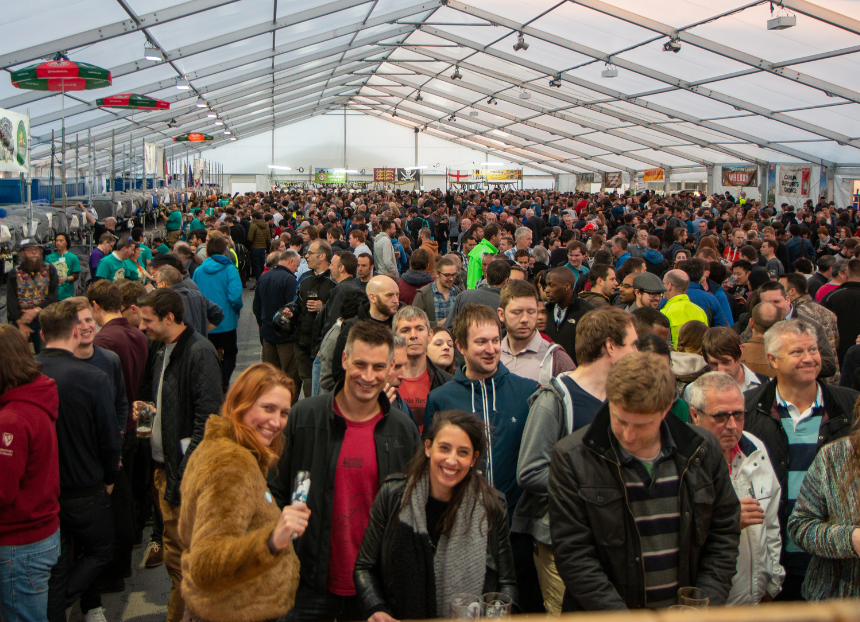 A bustling beer festival inside a large tent, with smiling attendees holding drinks, creating a lively and cheerful atmosphere.